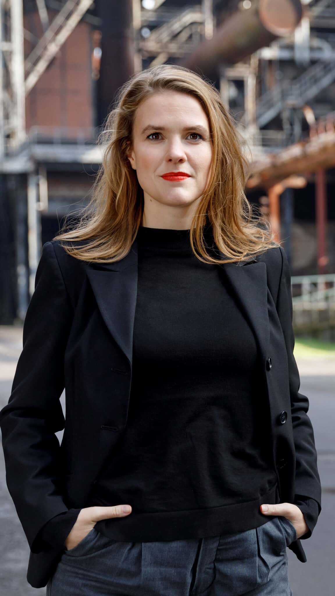 Terry steht vor einer Industriekulisse, sie hat die Hände in den Hosentaschen und schaut eine*n an.